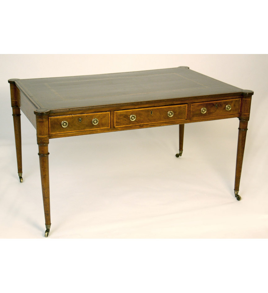 A late George III mahogany writing table, banded overall in satinwood, the leather top with turret corners, above three frieze drawers on the front and reverse, on ring-turned tapering legs, brass casters.  Handles replaced.