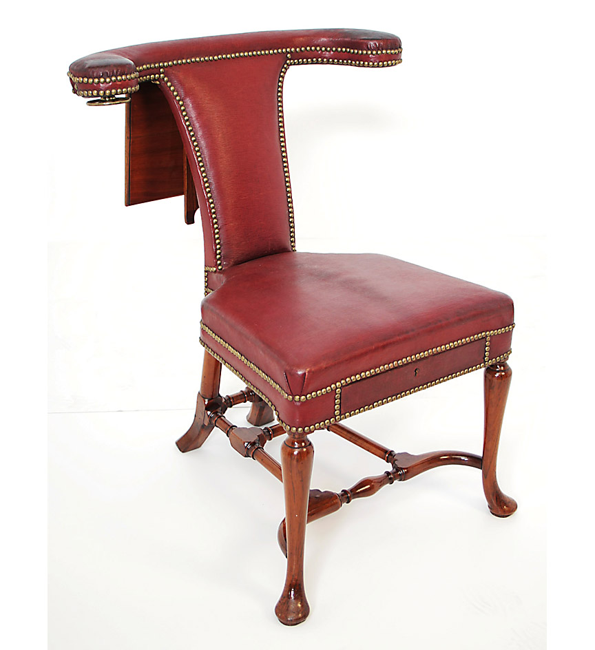 A rare Queen Anne walnut reading or 'cock fighting' chair upholstered in red leather, the horseshoe shaped back with the top rail terminating in hand rests, the original adjustable brass mounted swivel reading rest fitted with detachable brass candle arms; the arms fitted with a concealed inkwell and pen tray, a further drawer fitted to the seat rail, on splayed legs joined by an H-frame stretcher terminating in pad feet.