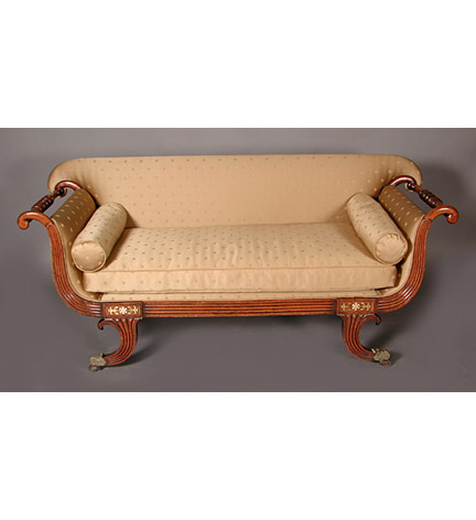 Regency upholstered and brass-mounted mahogany settee, the reeded seatrial continuing up to outscrolled arms with turned rails, and with splayed reeded legs ending in casters.  This settee is a Regency interpretation of the Grecian couch, with sinuous curves and scrolled ends.  See Hinckley's 