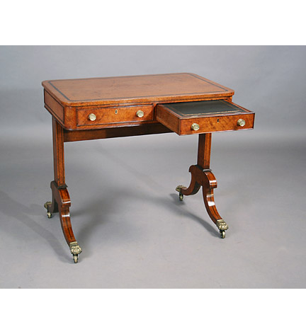 A small Regency inlaid amboyna trestle-end writing table, the top with ebony banding over a two-drawer frieze, one fitted with a writing slide, and raised on trestle supports ending in brass paw casters.