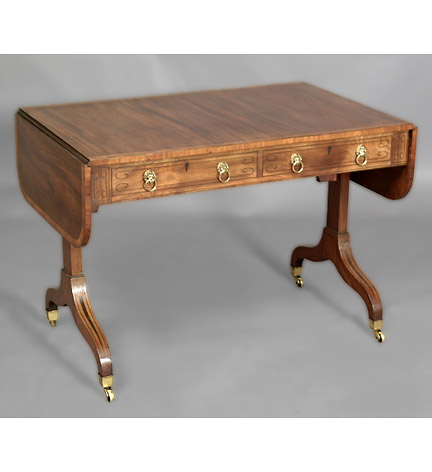 Regency mahogany and rosewood crossbanded sofa table, inlaid with boxwood and ebonized lines, the rounded with a pair of drop leaves above a pair of frieze drawers and opposing simulated drawer, raised on trestle supoprts with downswept legs ending in brass casters.
